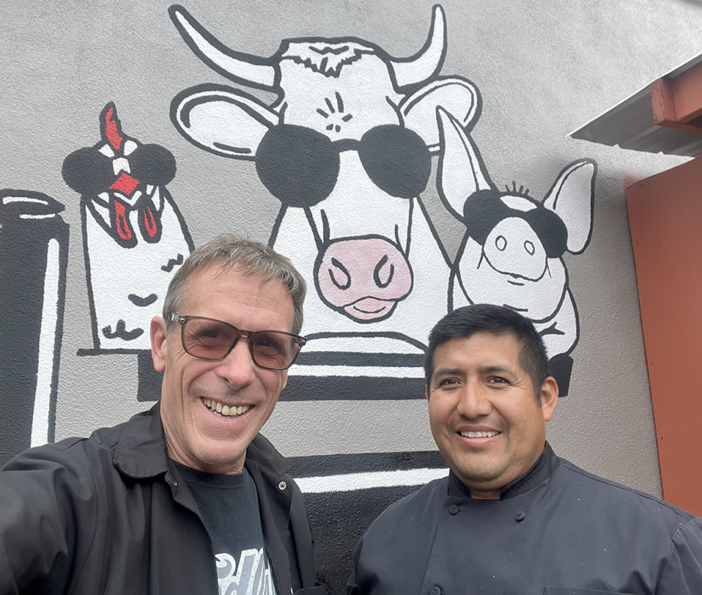 two men posing in front of a chicken, cow and pig mural