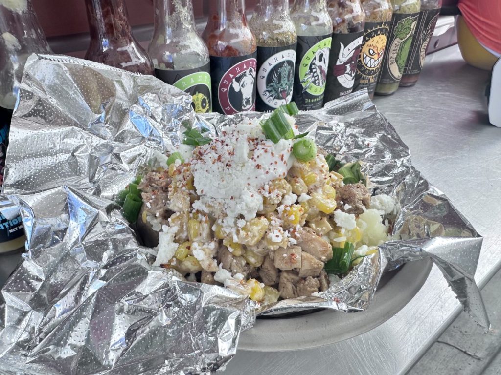 A baked potato filled with elote, creamy sauce, seasoned chicken, and sprinkled with cheese and Tajin.