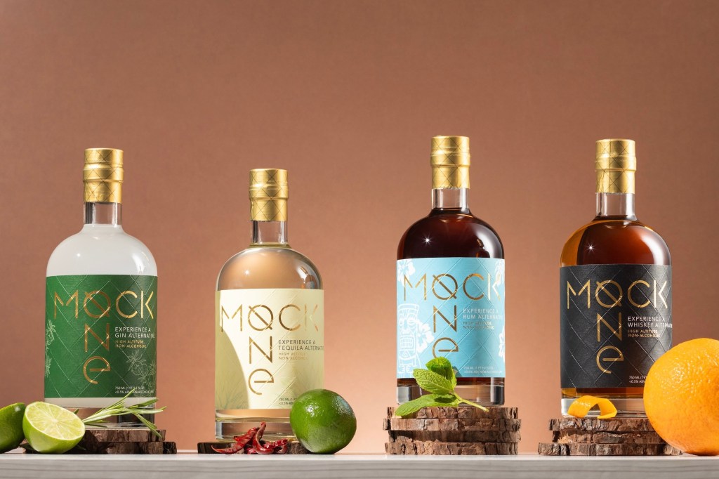 Four bottles of non-alcoholic spirits from Mock One set alongside fresh fruits and herbs