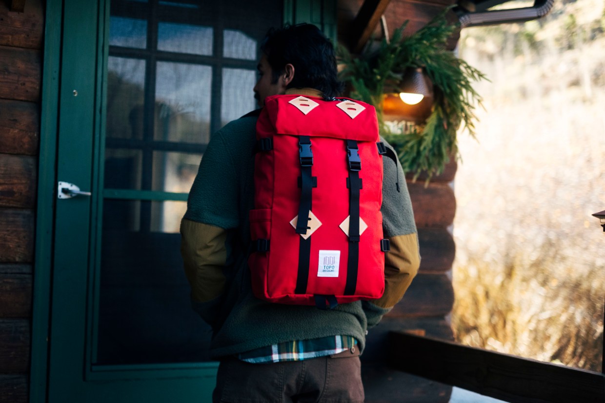 A person wearing a red Topo Designs backpack and apparel outside of a log cabin