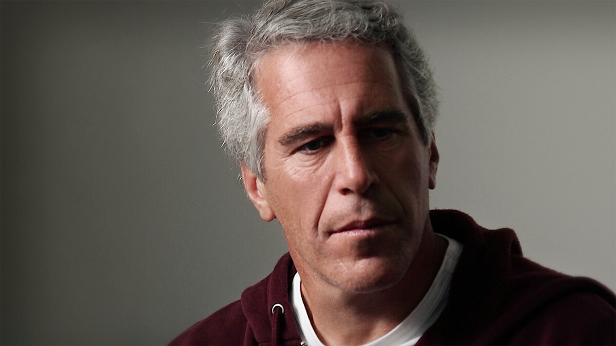 Jeffrey Epstein sits in front of a backdrop or grey all, looking down and away from the camera.