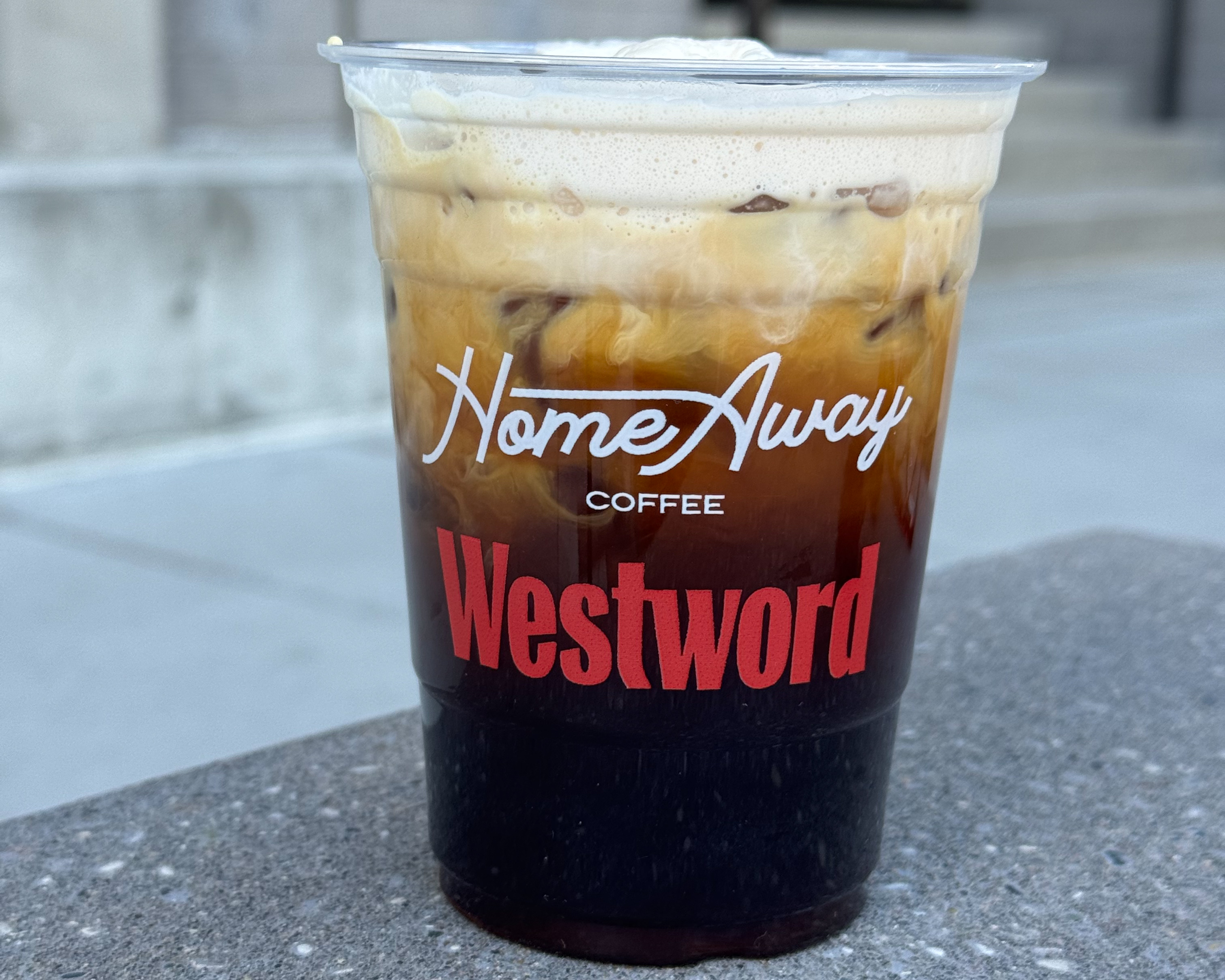 The new Westword cold brew drink with Home Away Coffee sitting on a table outside. The cup has the Home Away Coffee and Westword logos.