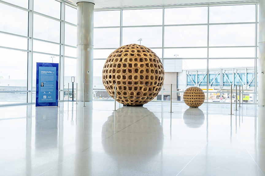 Two large wooden spheres with a perforated design are displayed in a bright, spacious airport terminal.