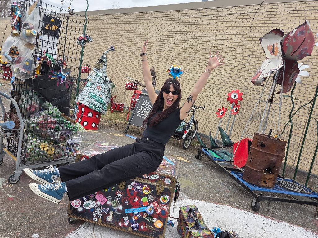 Cass Ivey sits on a hard-case gear box, legs outstretched and arms in the air, grinning.
