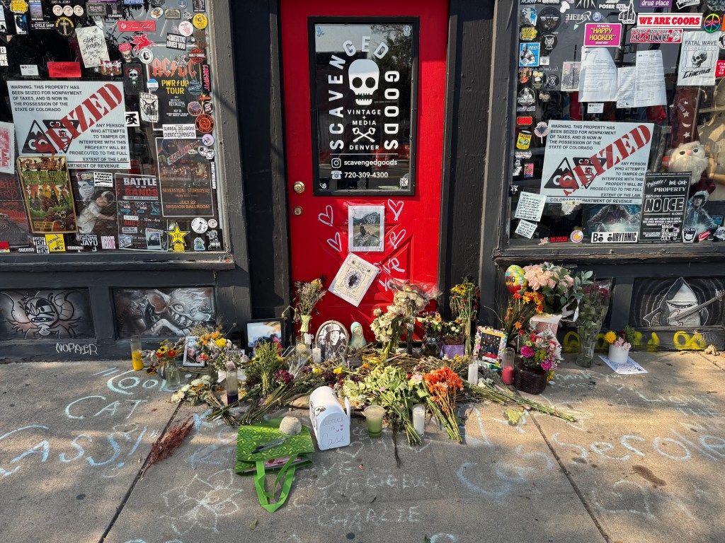 A memorial for Cass Ivey on the sidewalk in front of Scavenged Goods, featuring flowers, photos, and other offerings.