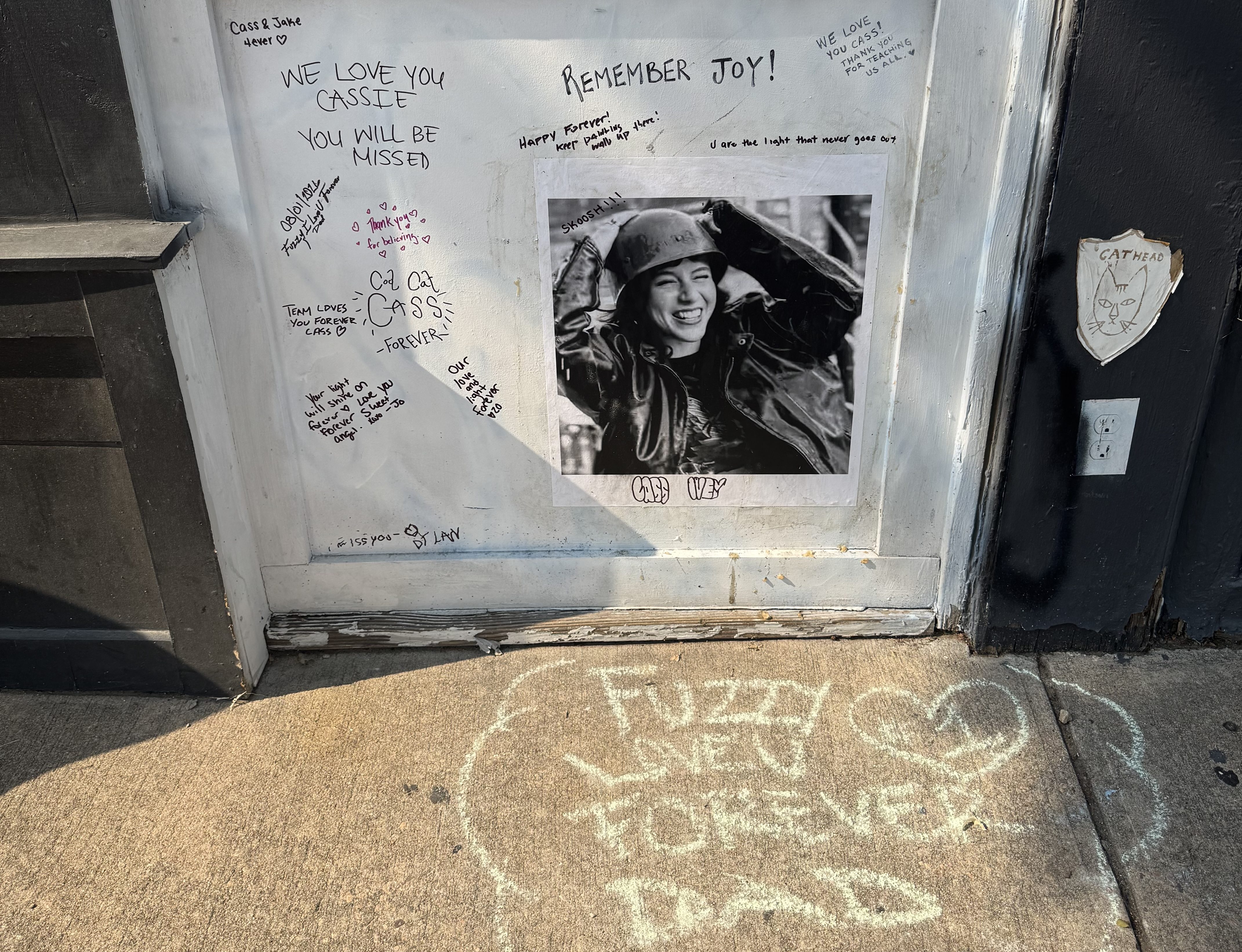 A white wall with handwritten notes to Cass and a photo of her smiling. On the sidewalk, a note in chalk reads "Fuzzy, love forever, Dad" with a #1 in a heart.