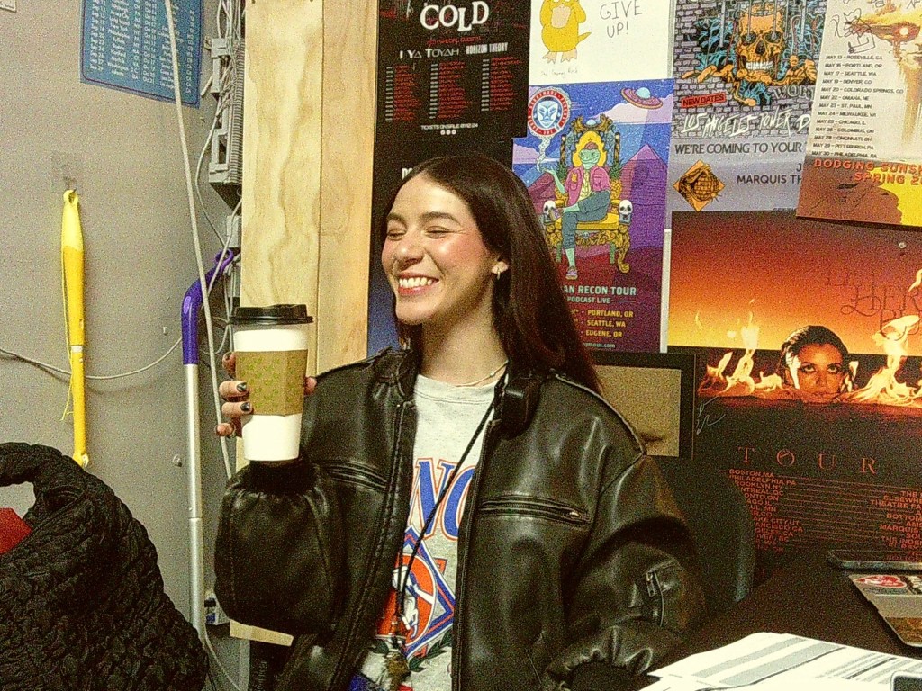 Cass Ivey grins and holds a large to-go cup. Concert posters cover the wall behind her.
