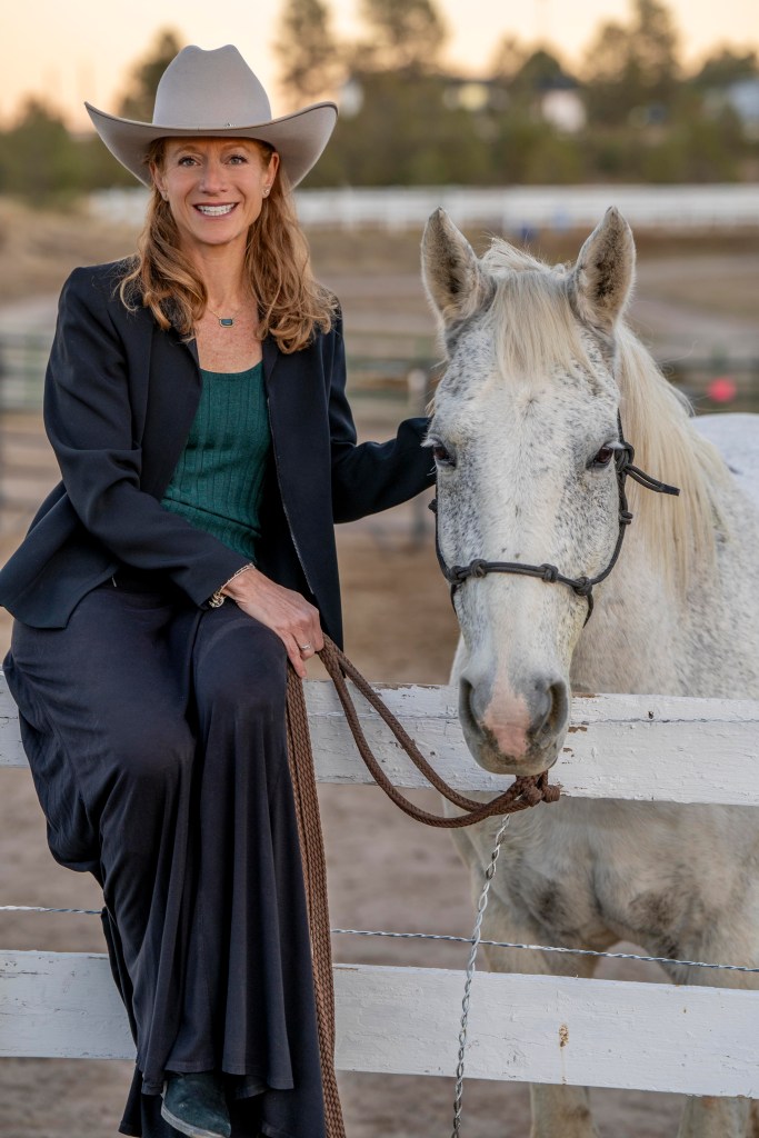 Kaye in a Stetson hat, seated on a white fence next to a horse