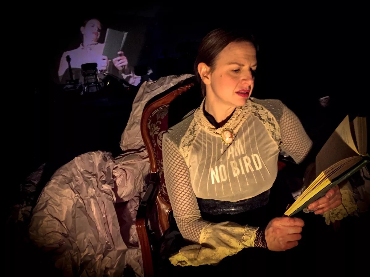 A woman sits in an old-fashioned chair, reading a book, while her image is projected behind her.