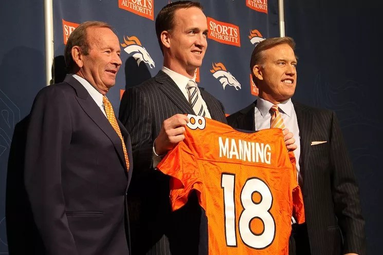 Three men stand in a row, one holding up an orange jersey.