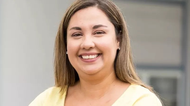 A woman in a yellow shirt smiles widely at the camera. She is pictured from the shoulders up.