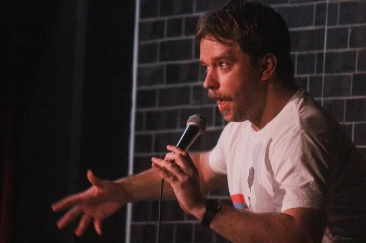 A man with a mustache speaks into a microphone on stage.