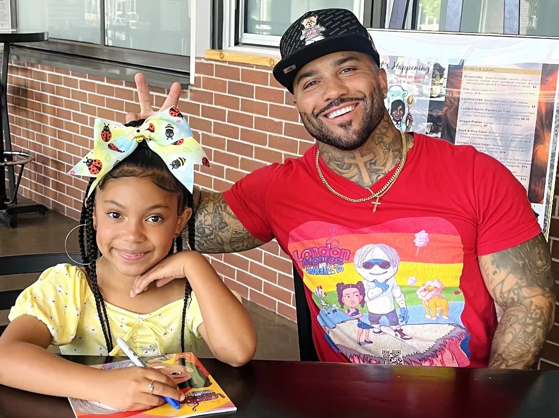 Girl signing colorful children's book sits to the right of father, who is holding two fingers above her head.