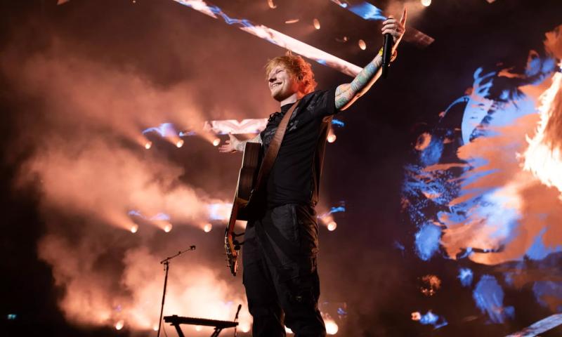 red head stands on stage with his arms held open for applause