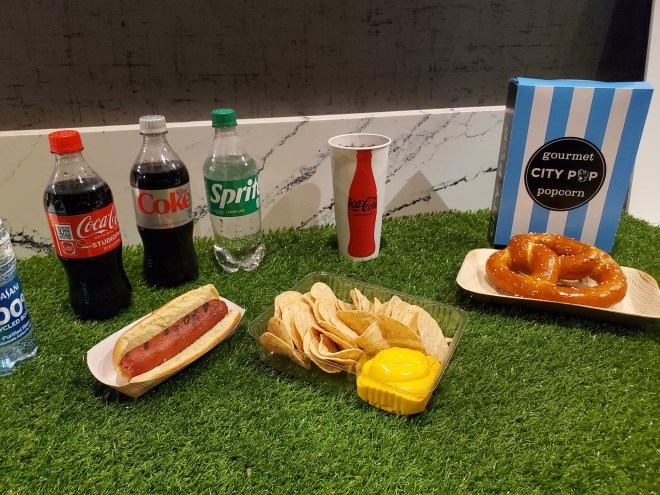 bottles of soda, a hot dog, nachos, a pretzel and a bag of popcorn