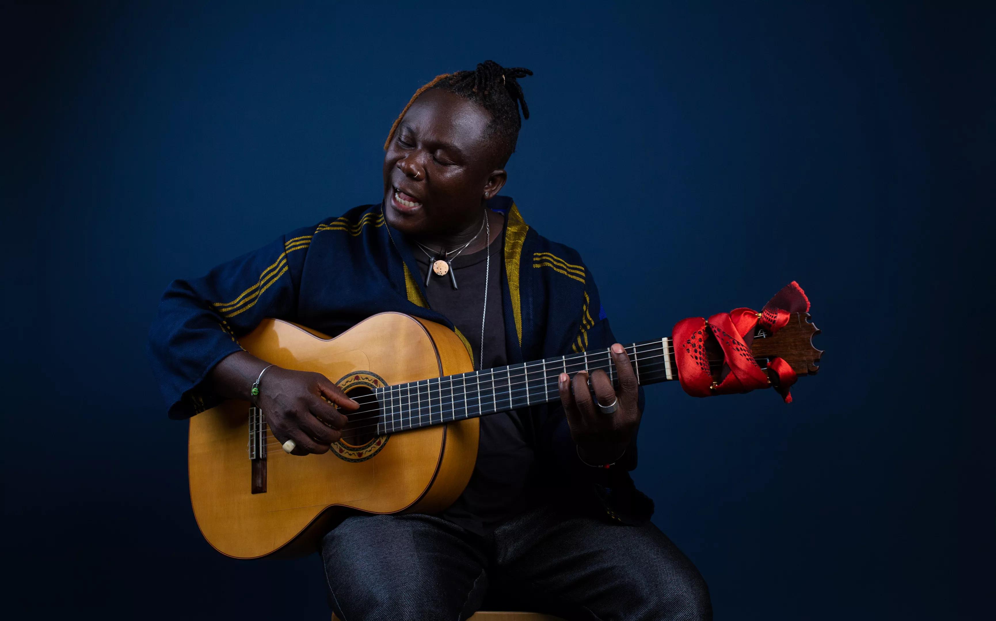 Seated man singing with guitar on blue background.