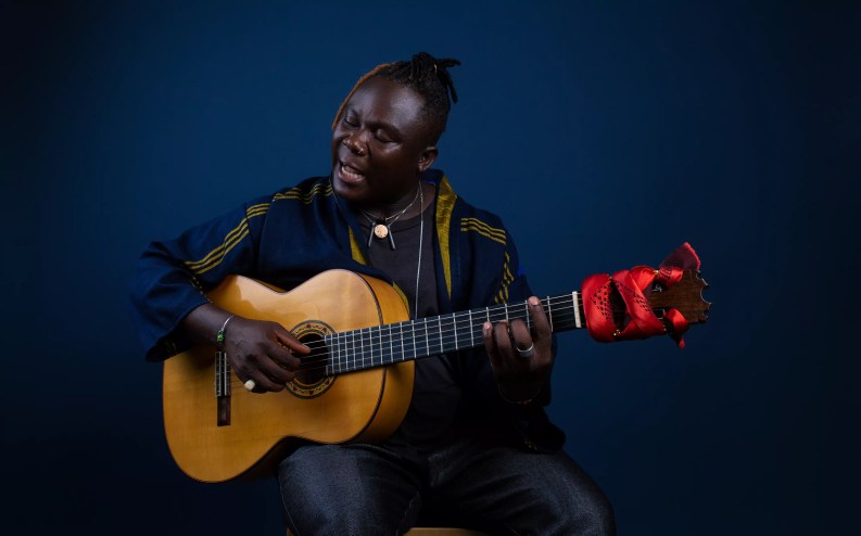 Seated man singing with guitar on blue background.