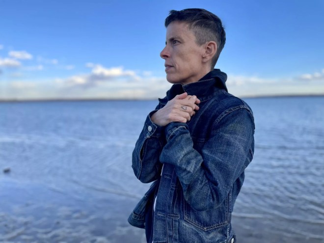 person with short hair has hands to the heart as they stand in front of water
