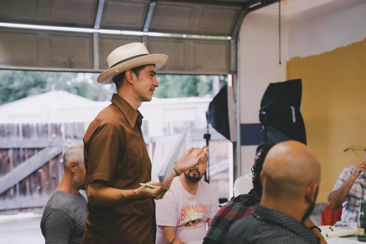 man in white hat and brown shirt speaks to other seated people