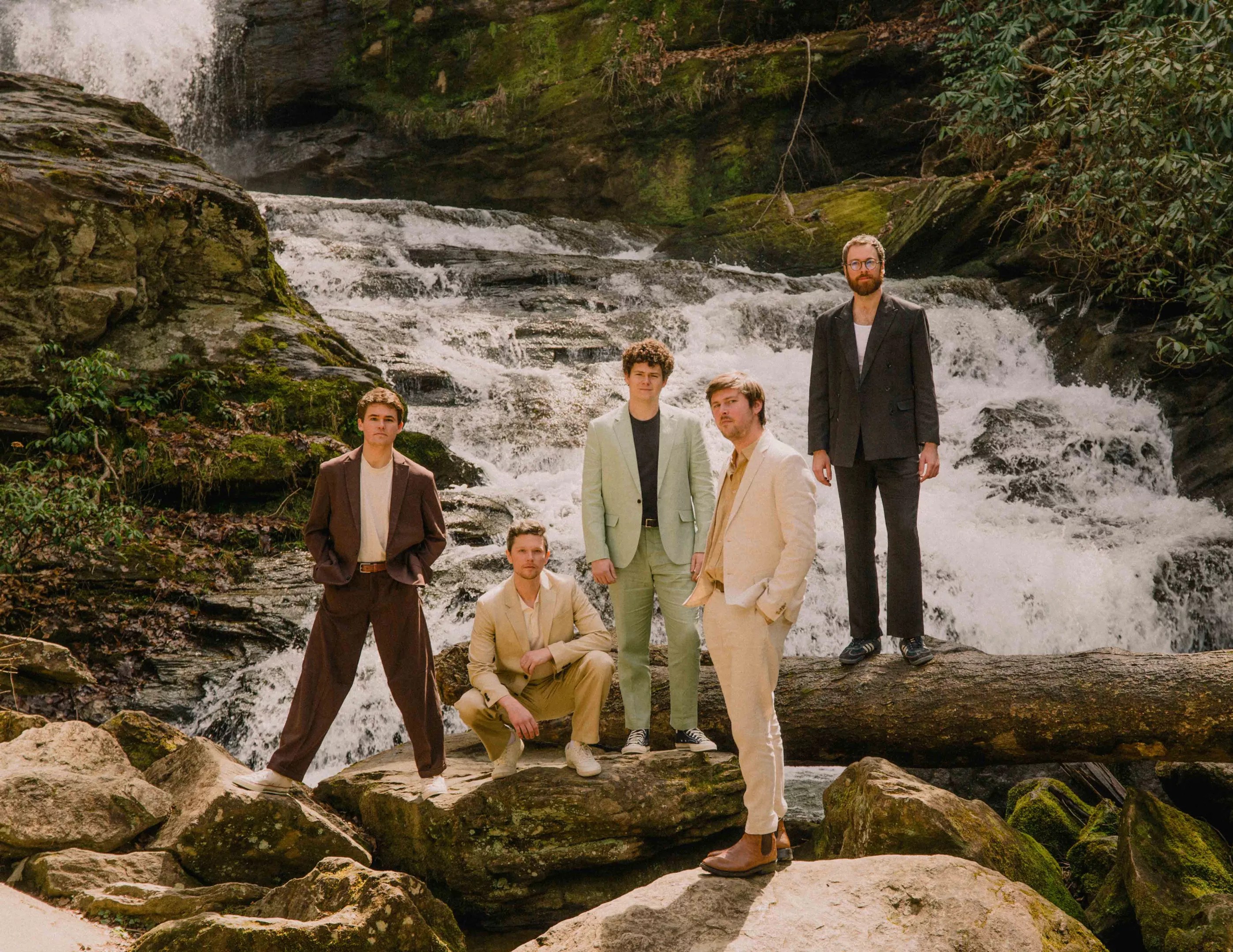 Four men standing, one squatting, on top of rocks and logs above a running creek. All dressed in different color suits.
