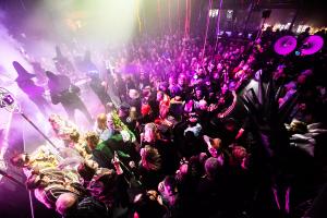 a crowd dances in a concert venue to masked drummers on stage wearing witch hats
