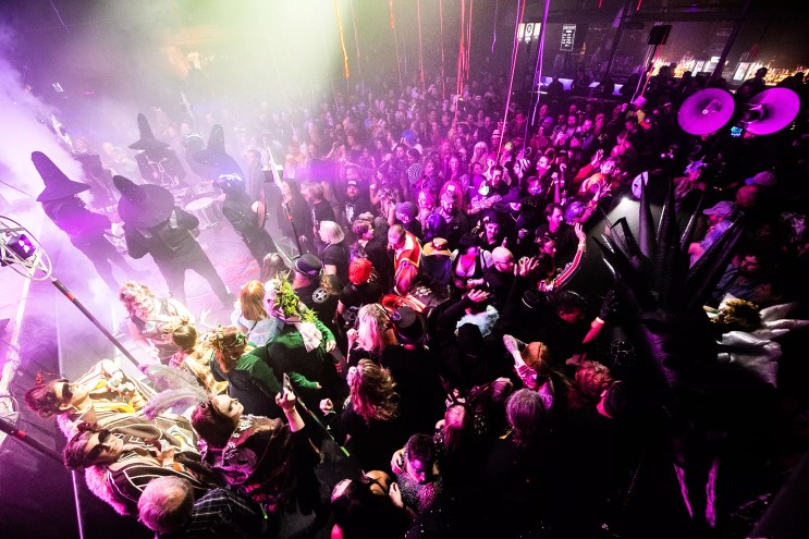 a crowd dances in a concert venue to masked drummers on stage wearing witch hats