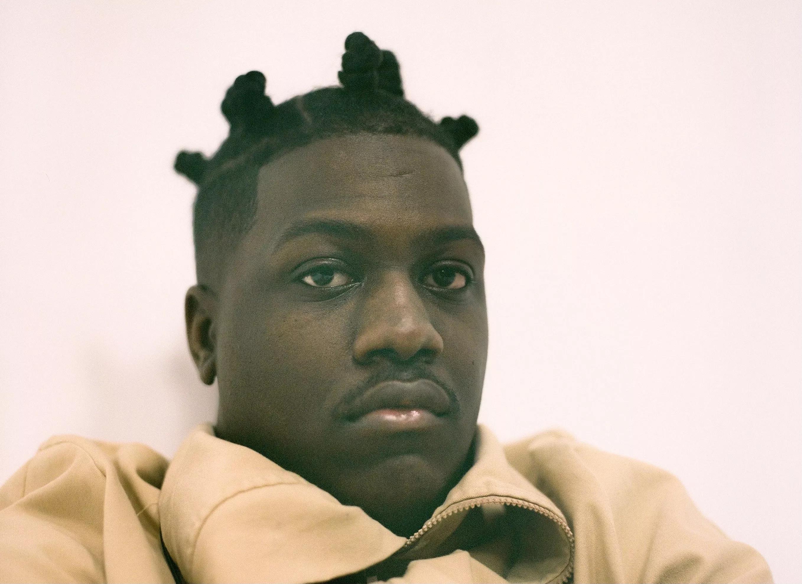 black man in an orange sweater sitting against a pink wall