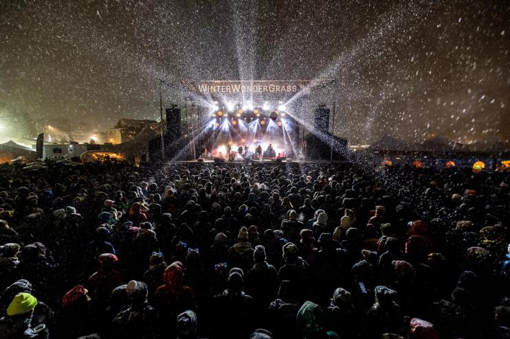 a stage with the words WinterWonderGrass is surrounded by an audience in the snow