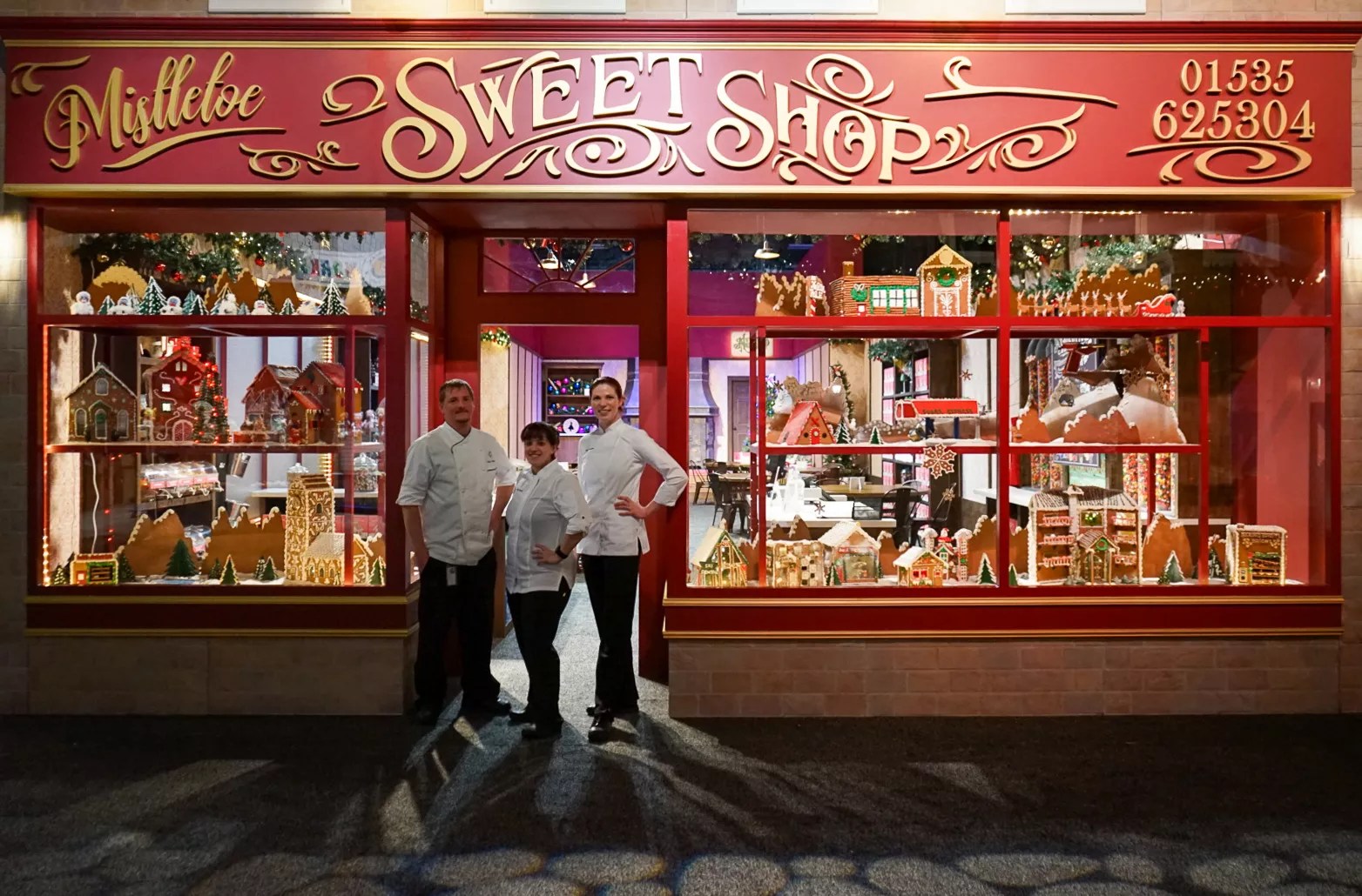 three people in chef's coats standing in front of a gingerbread display in a store window