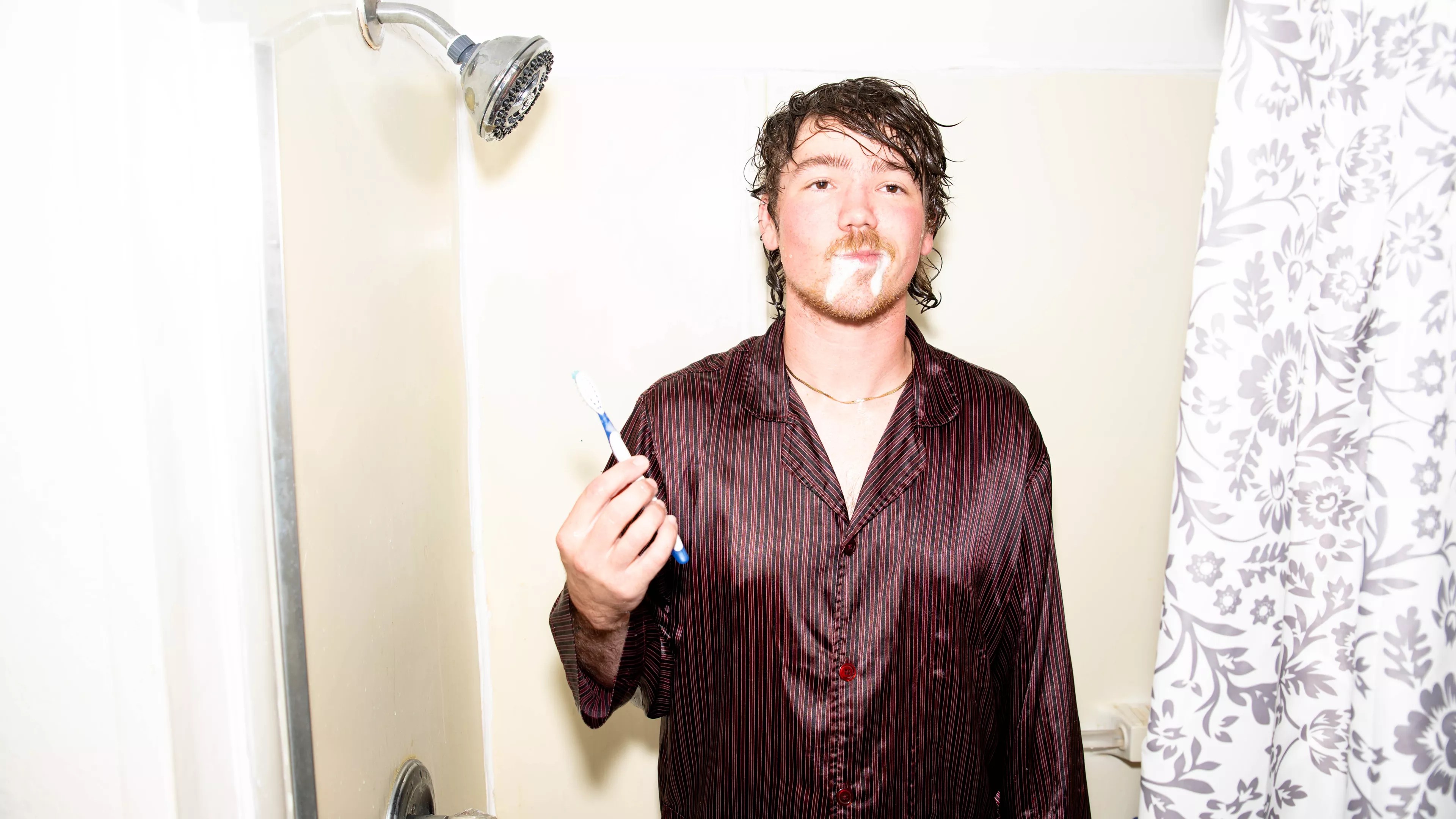 man brushes teeth in the shower wearing a brown silk shirt