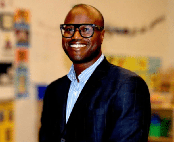 A man smiling in glasses and a blazer with a collared shirt.