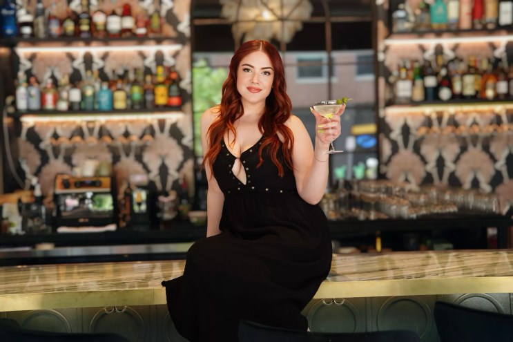 woman n a black dress sitting on a bar and holding a martini