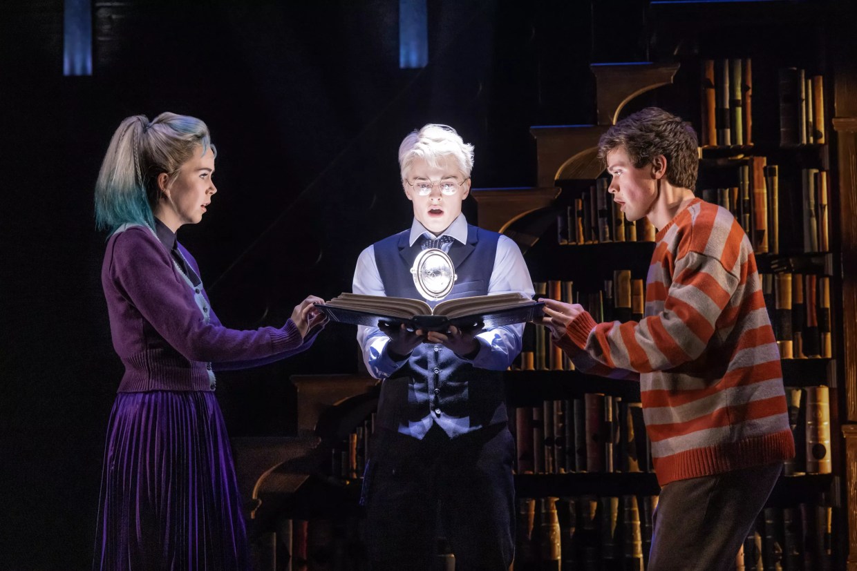 A man with silver hair holds a book, which two people grasp on either side.