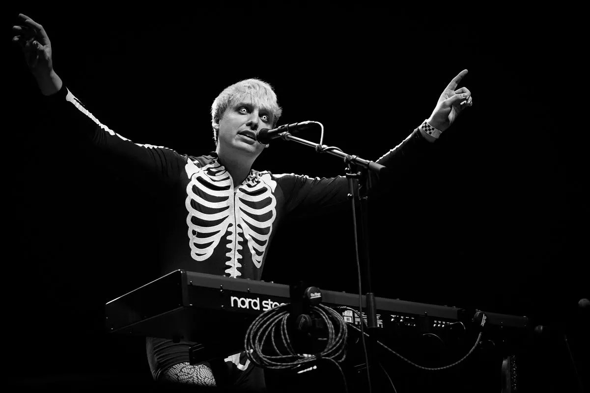 man in skeleton sweater stands at a keyboard and mic with arms in the air