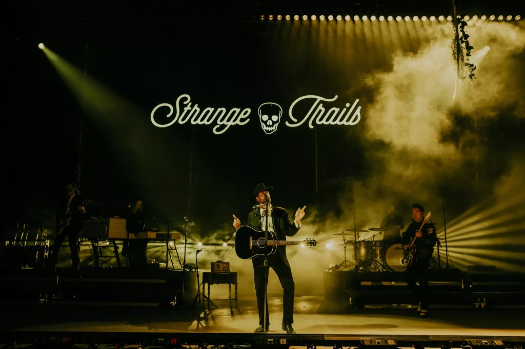Lord Huron performing on stage at Red Rocks with fog behind him, and a stage sign labeled Strange Trails hangs above him.