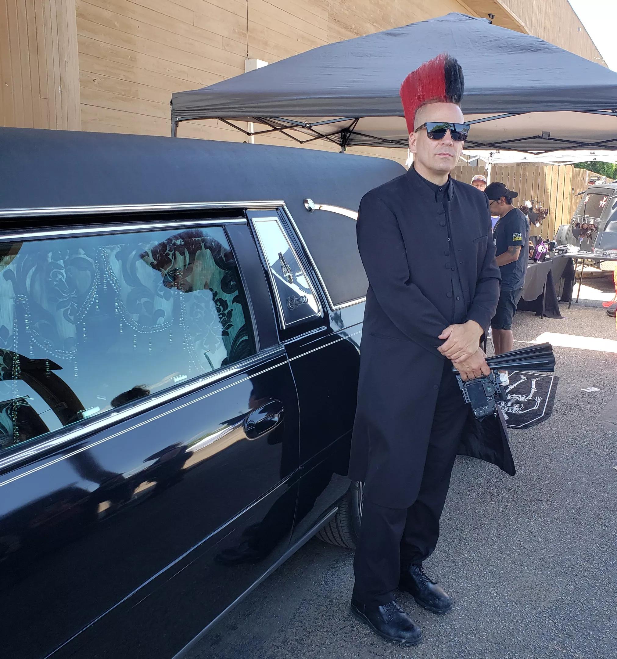 A man with a mohawk stands next to a hearse