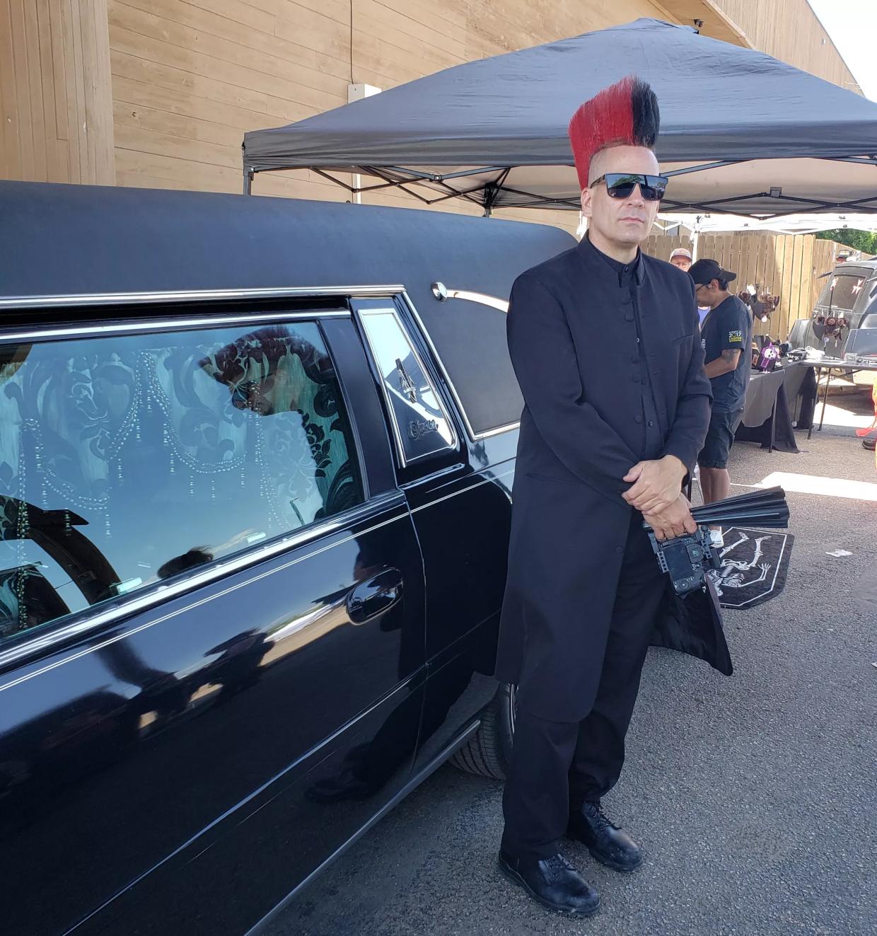 A man with a mohawk stands next to a hearse