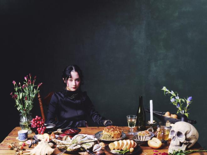 Someone sitting at a dinner table with a skull and several food items on a dark green-blue background